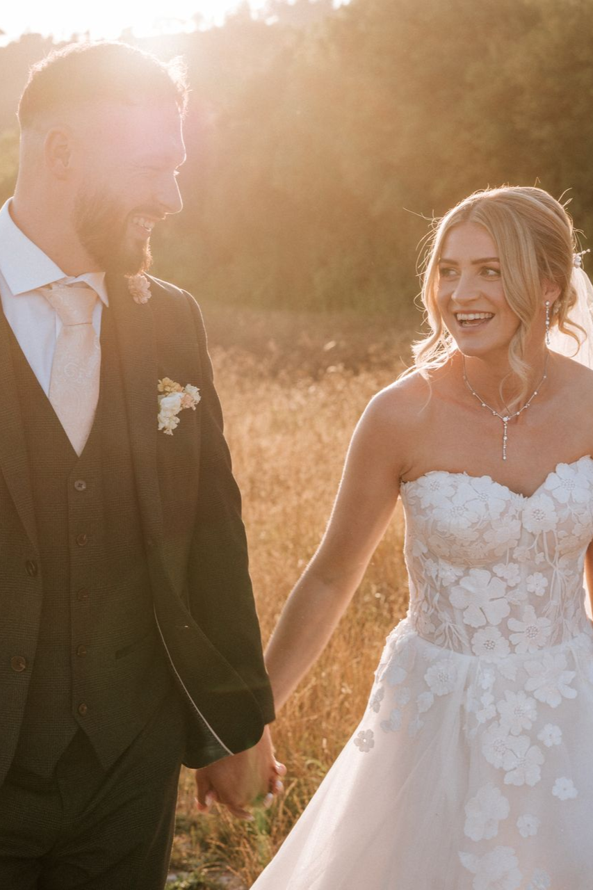 Bride and groom walking hand in hand at sunset, smiling in a sunlit field