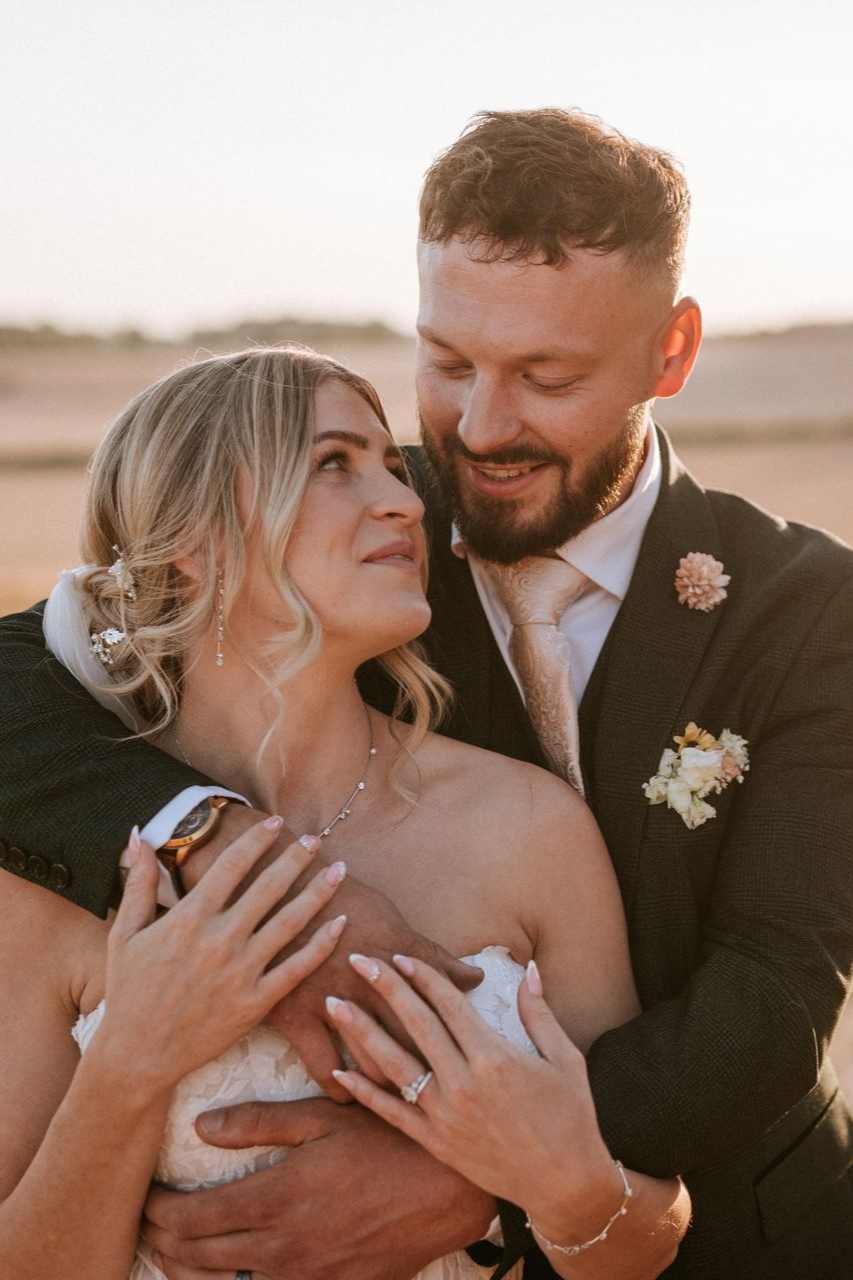 Couple embracing outdoors at sunset, smiling in wedding attire
