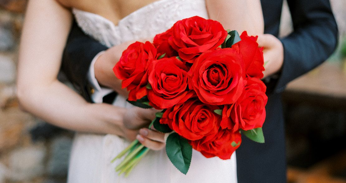 Bride holding a bouquet of red roses beside a groom in a wedding setting