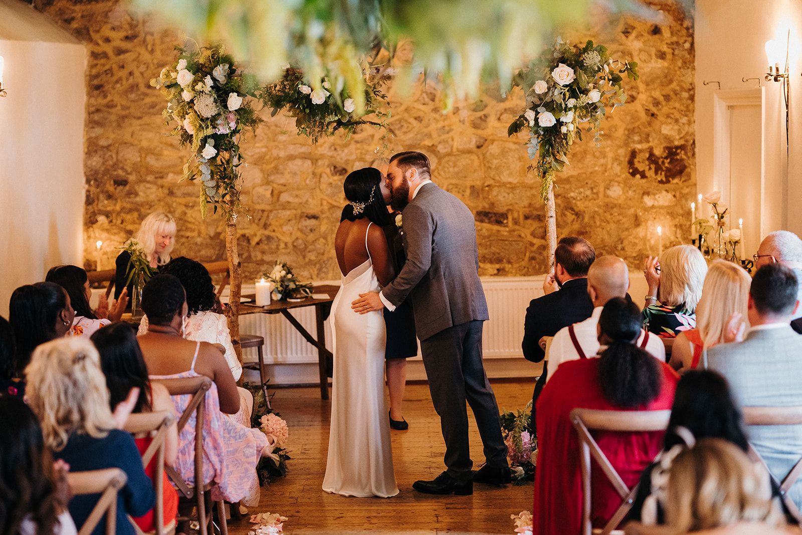 Wedding couple kissing at the altar, with seated guests and floral arch decor in a warm indoor venue
