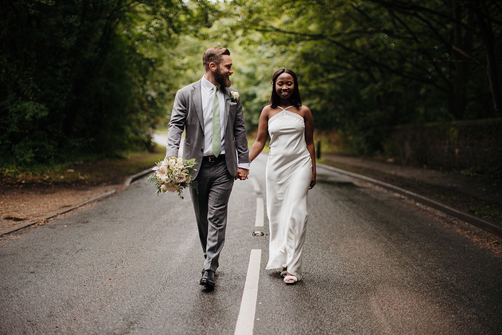 Bride and groom walking hand in hand on a wet tree-lined road, smiling and carrying a bouquet