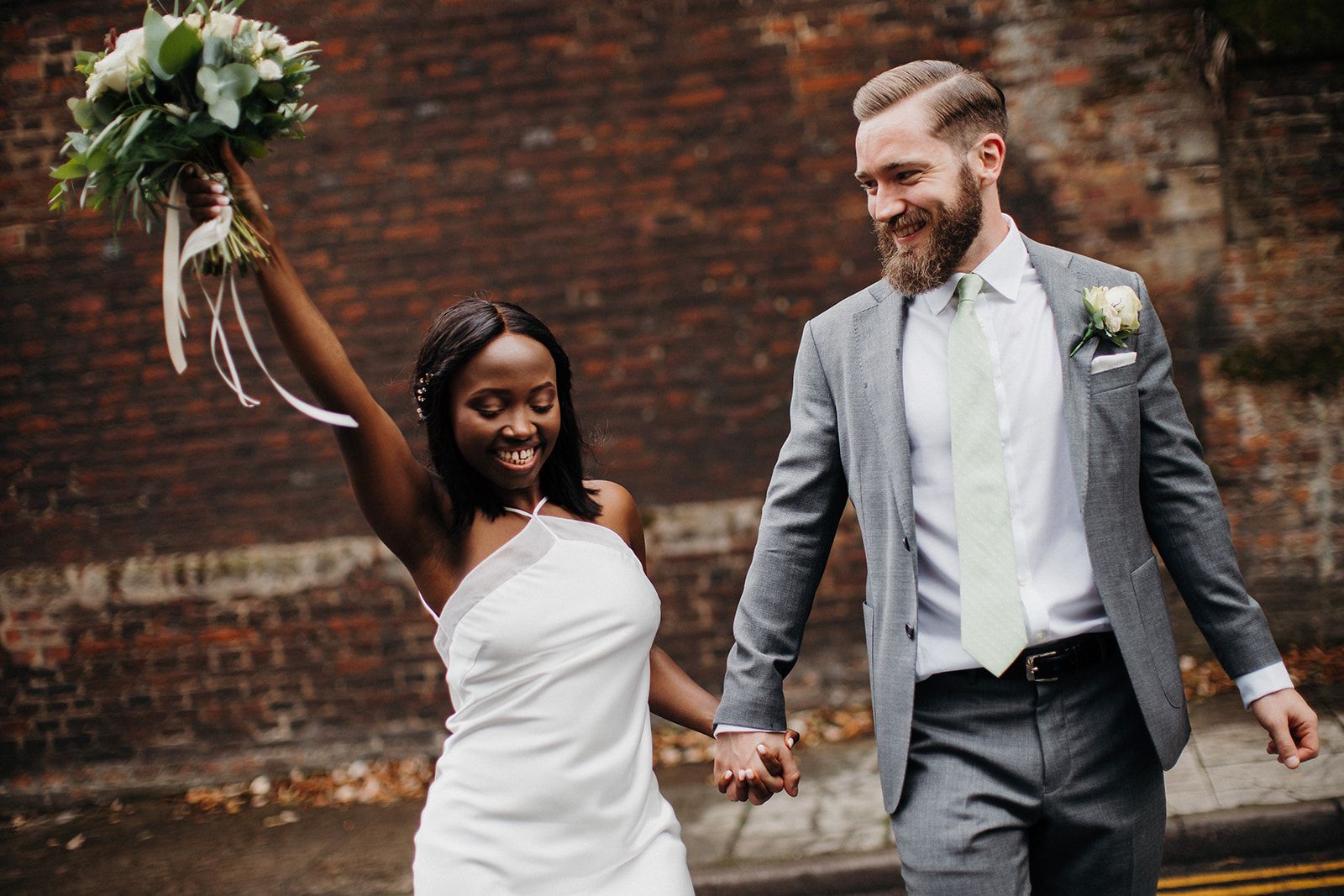 Bride in white dress raising bouquet while walking hand-in-hand with groom in gray suit outdoors