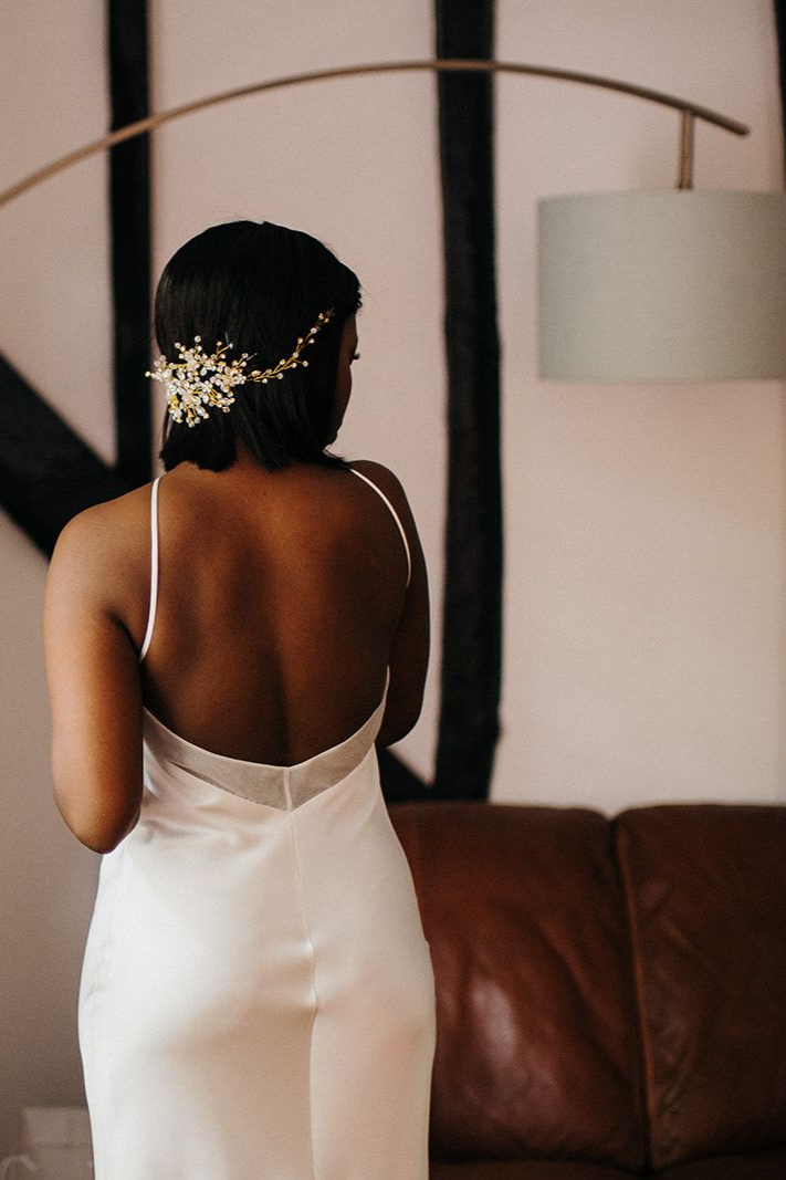 Woman in a white satin dress standing by a brown leather couch, seen from behind with floral hair accessory.
