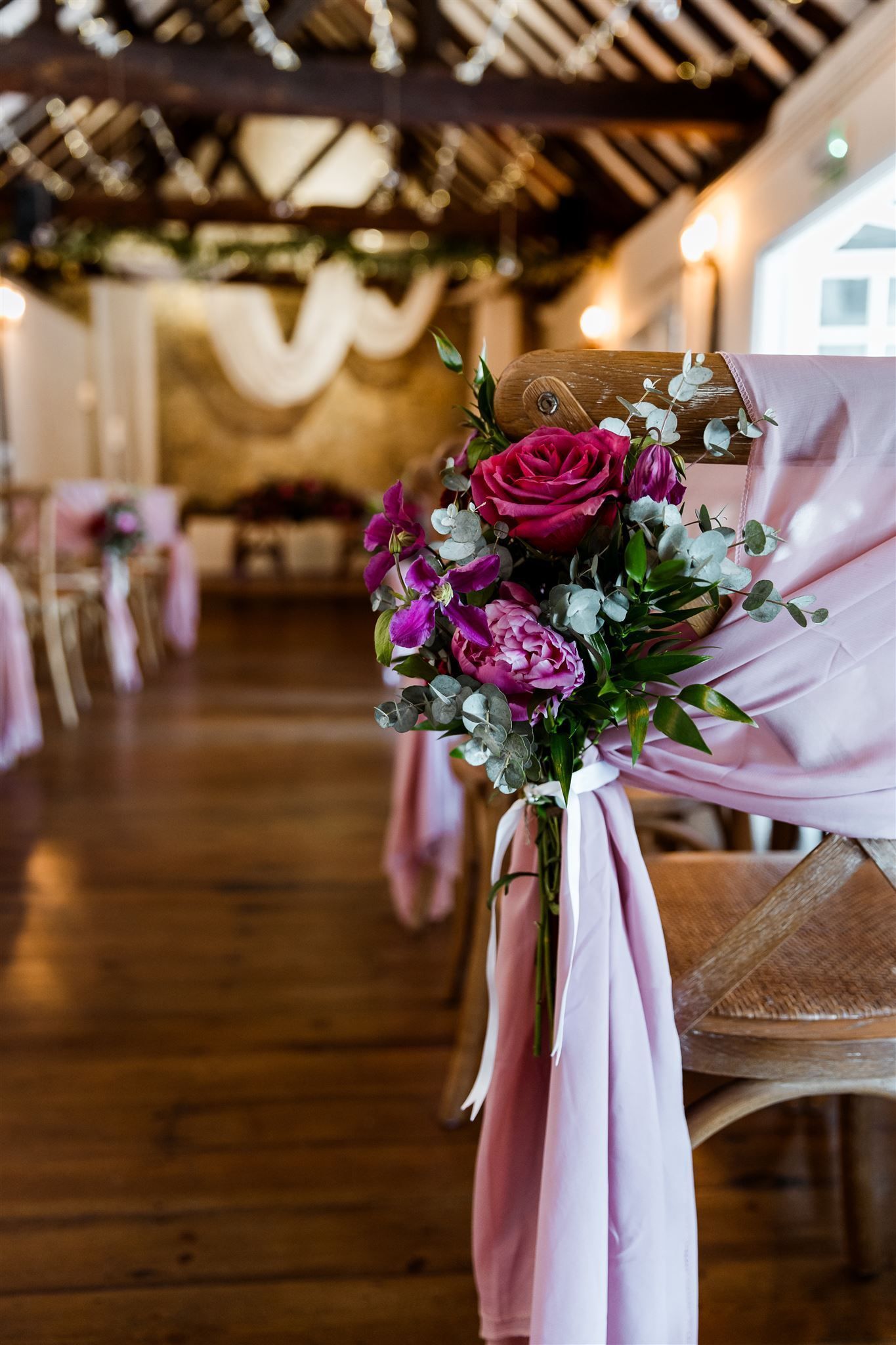 Rustic wedding venue with pink-draped chairs and purple floral bouquets along a wooden aisle