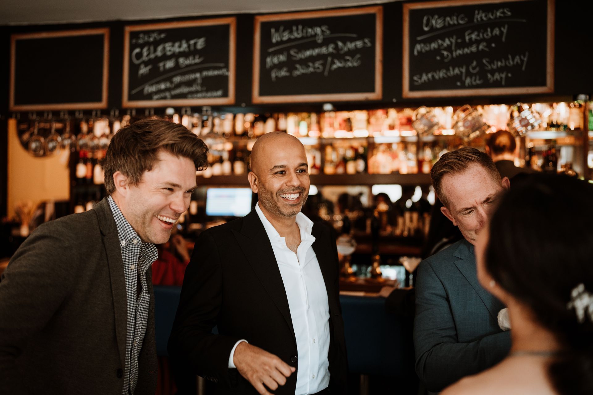 Three people chatting and smiling in a warmly lit bar with chalkboard menus behind them
