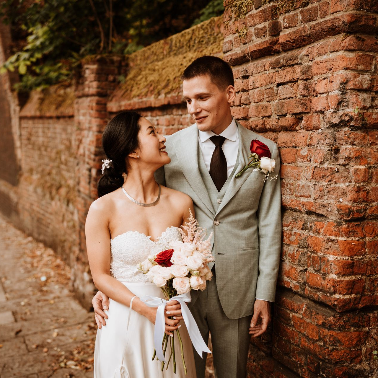 Bride and groom smiling beside a brick wall, the bride holding a bouquet