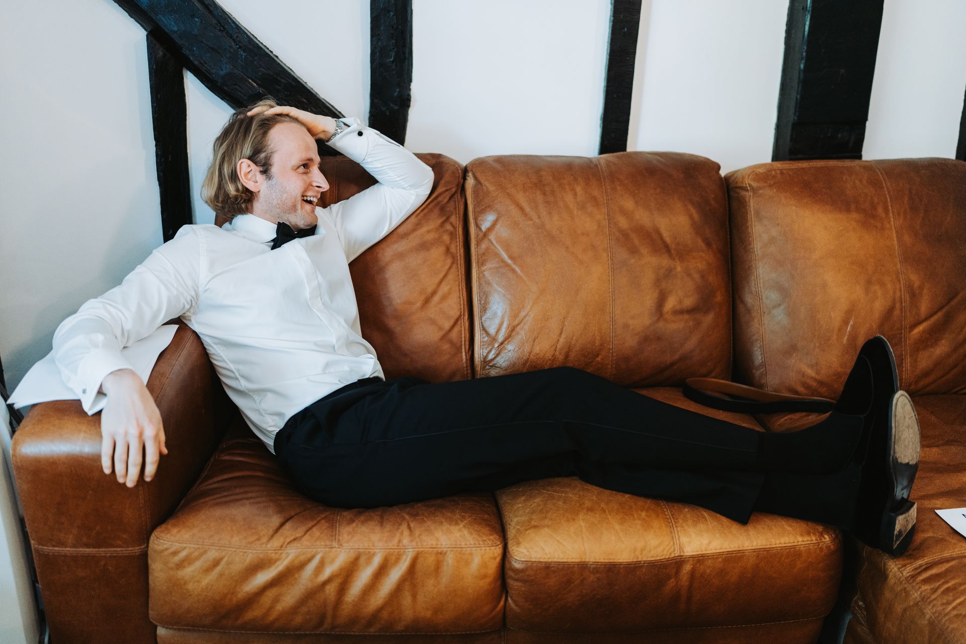 Person reclining on a brown leather couch in a bright room with black beams.