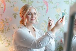 Woman applying makeup while looking into a hand mirror in a pastel floral room