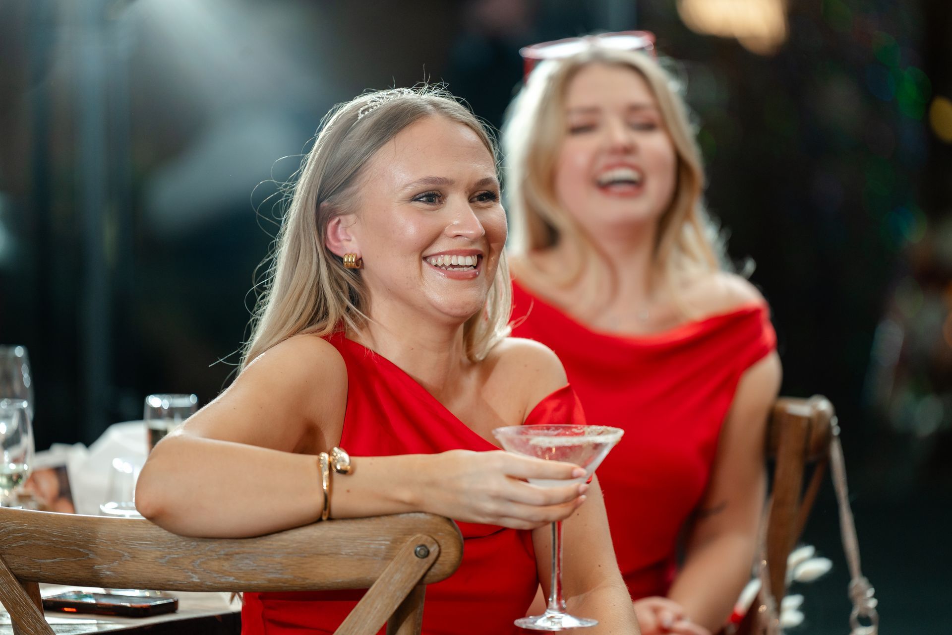 Two women in red dresses smiling at a table, one holding a martini glass.
