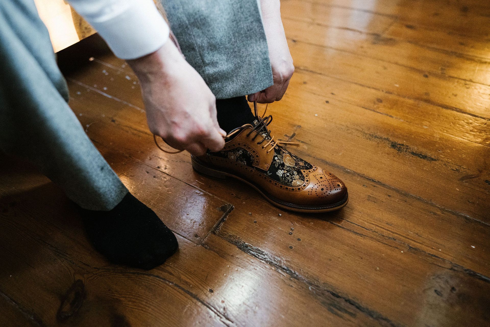 Hands tying a brown dress shoe on a wooden floor, with a black shoe nearby