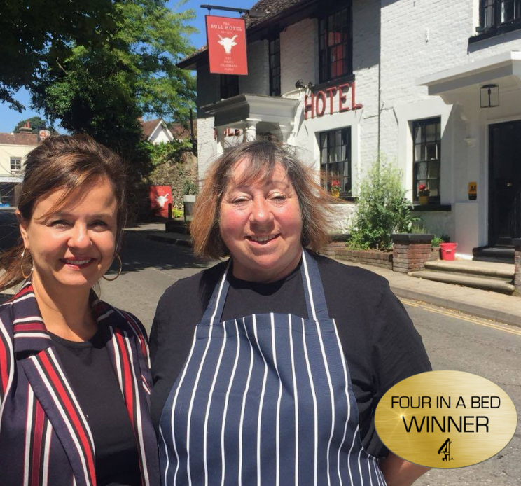 Two women stand outside a hotel, smiling, with a “Four in a Bed Winner” sign in the corner.