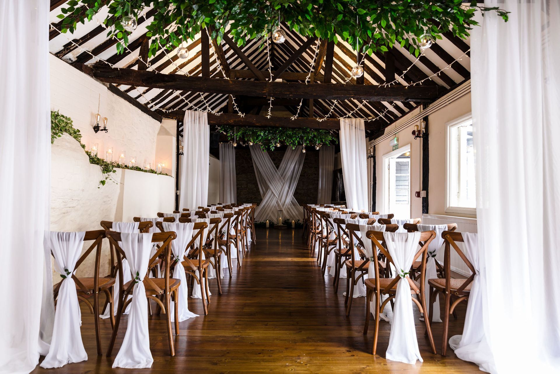 Elegant outdoor wedding aisle with white drapes, wooden chairs, and greenery overhead