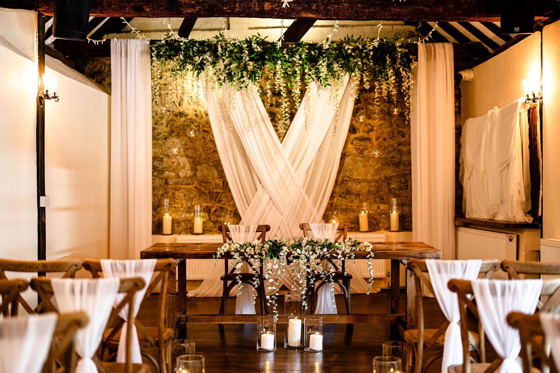 Rustic indoor wedding reception aisle with wooden chairs, white drapes, and greenery overhead
