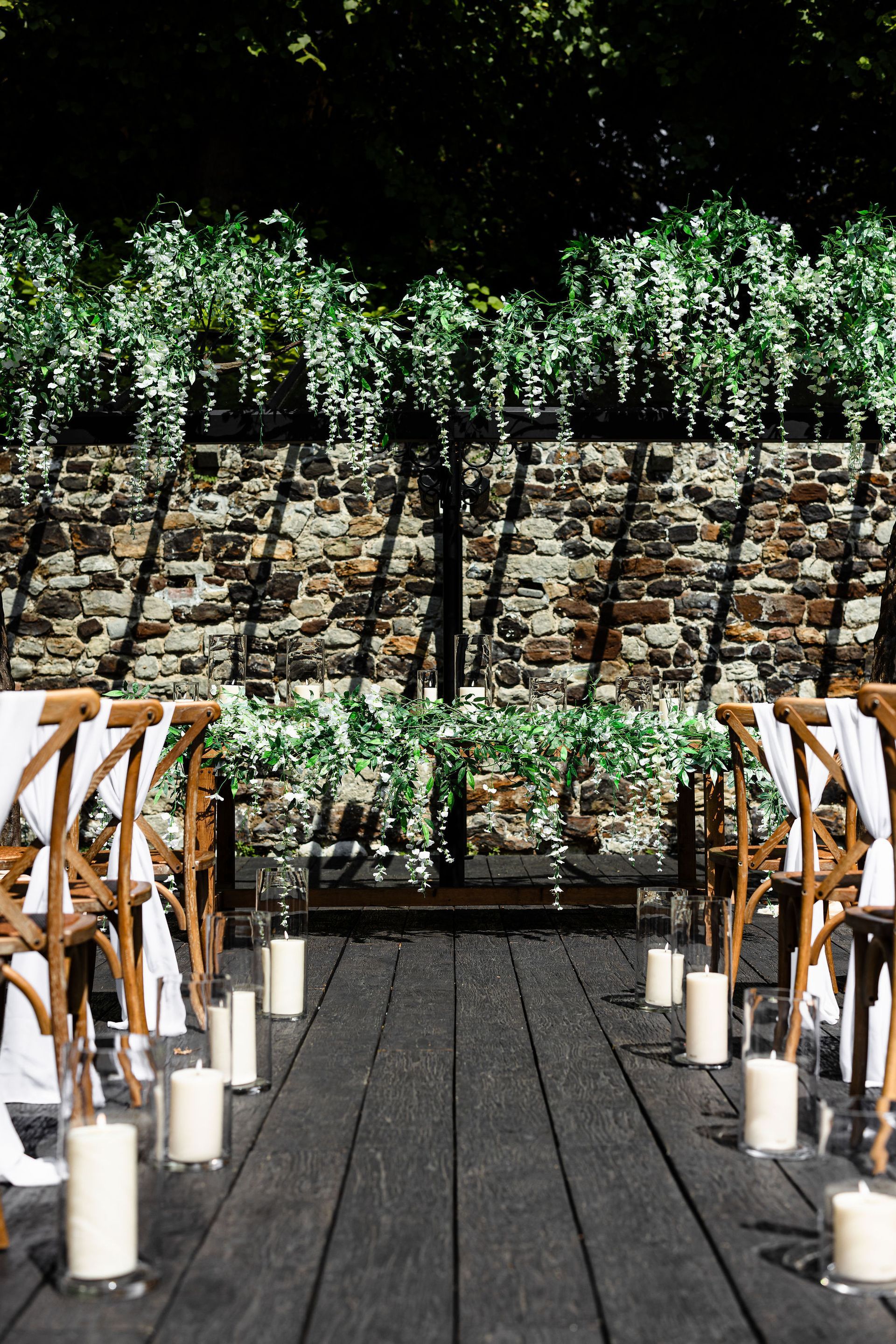 Outdoor wedding aisle with white candles, wooden chairs, and a floral arch backdrop