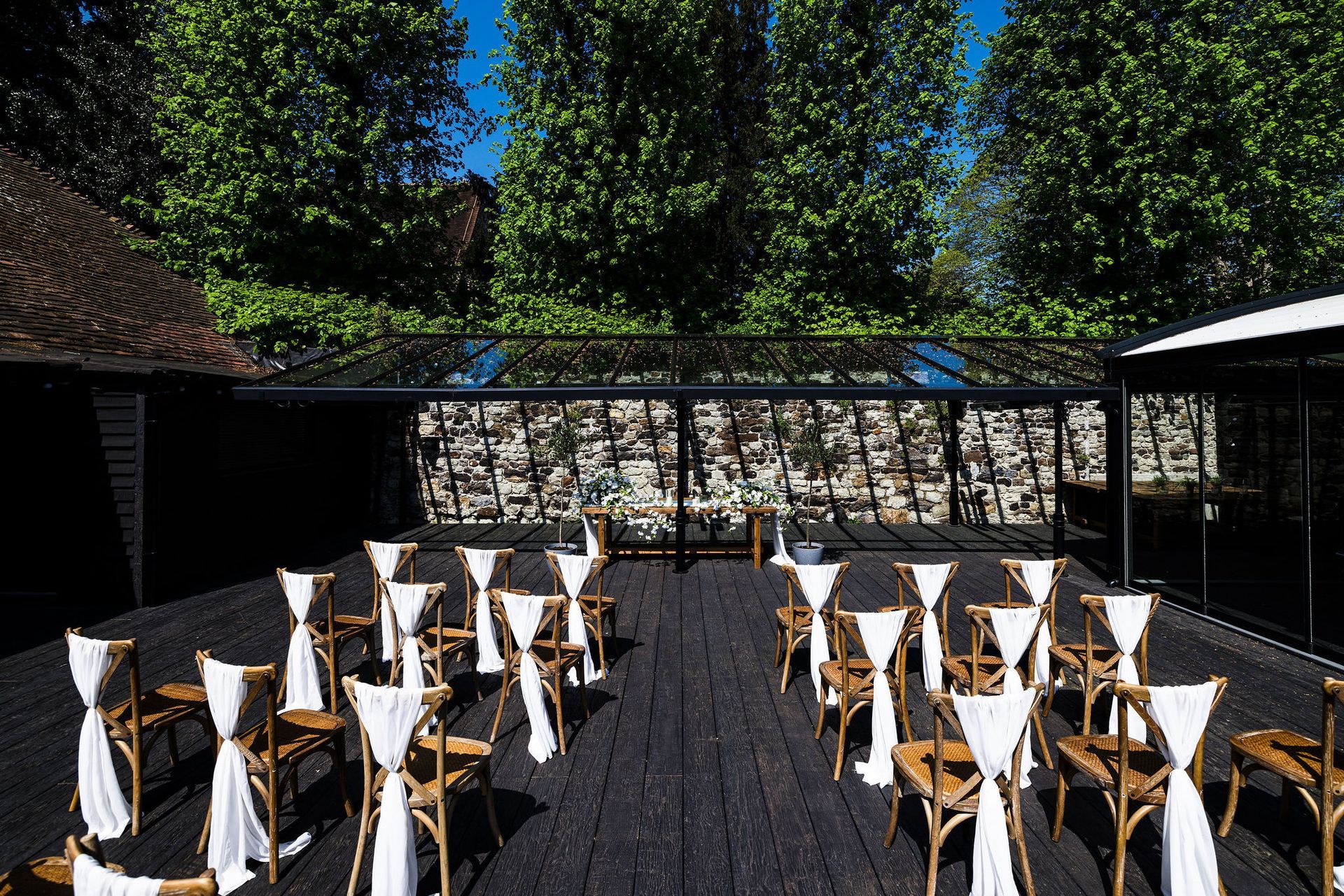 Outdoor café with seated people in a sunny courtyard beside a brick building and glass terrace.