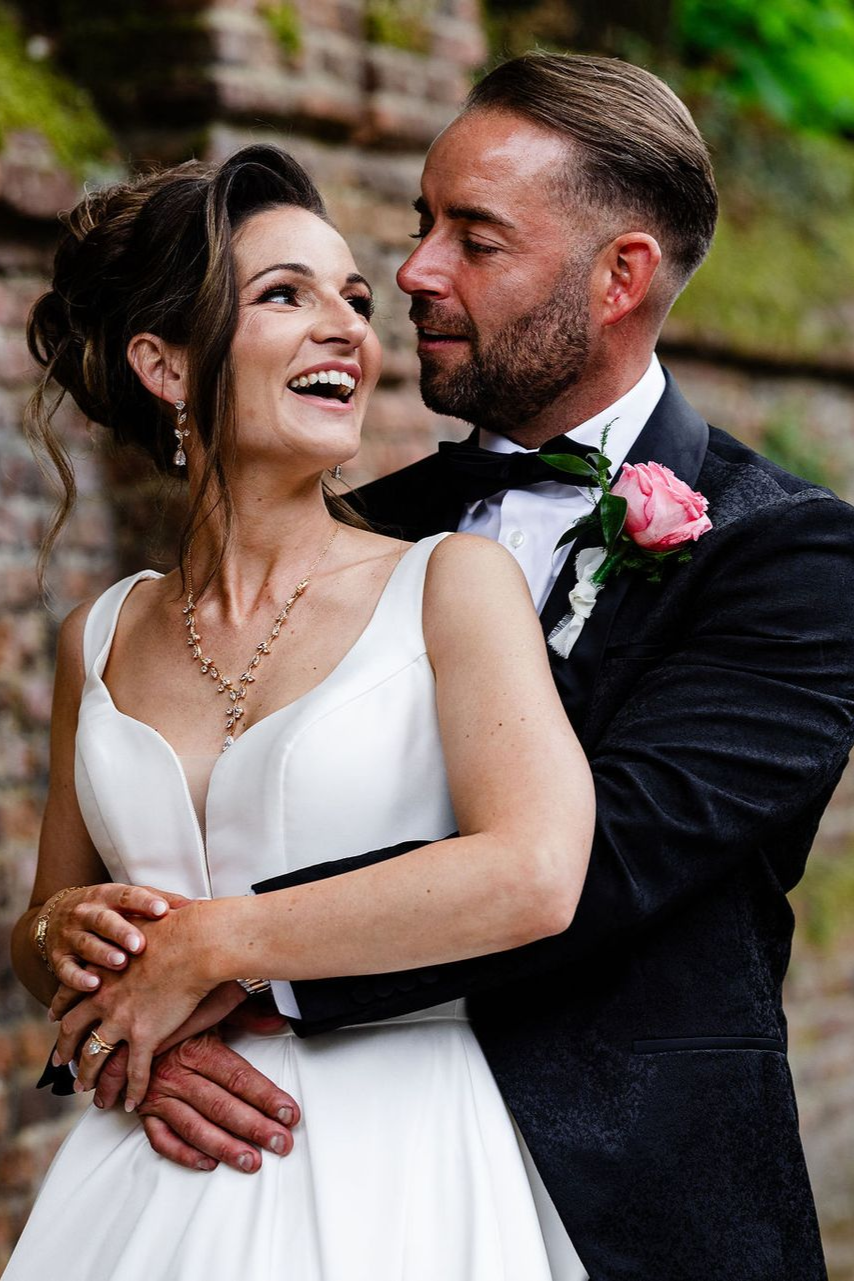 Smiling bride in a white dress embracing groom in a dark suit with pink boutonniere outdoors