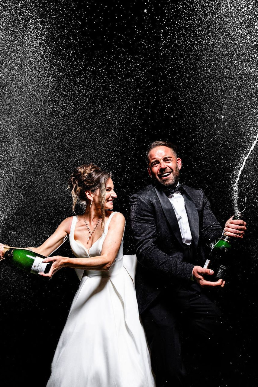 Bride and groom pop champagne in celebration amid falling confetti, smiling in formal wear