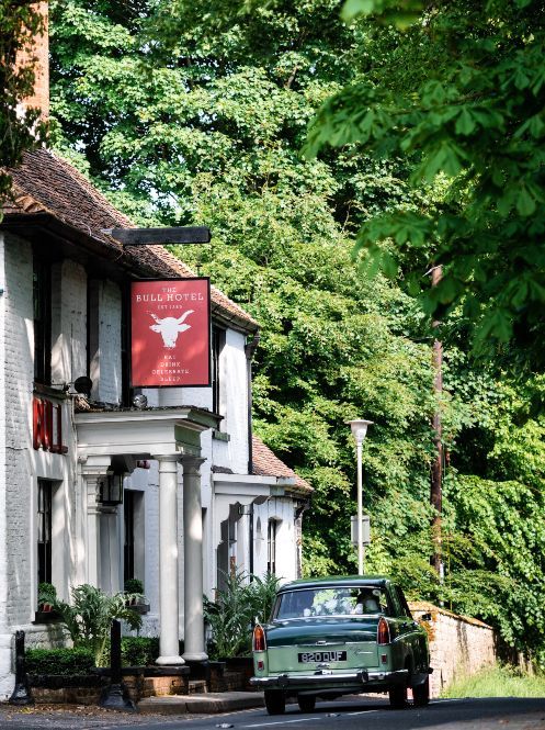 Black vintage convertible parked outside a white inn on a tree-lined road with a red sign.