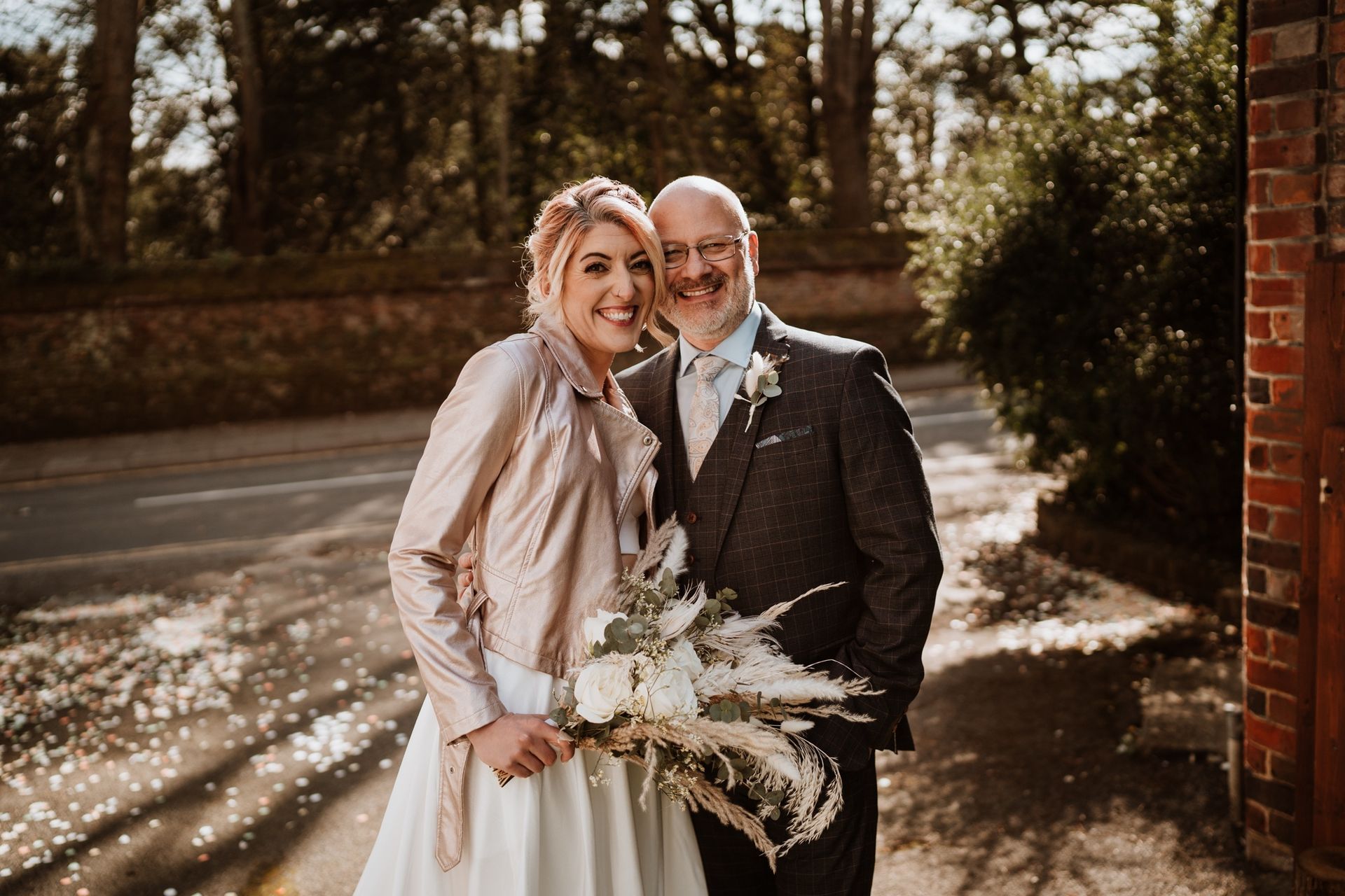 Smiling couple in wedding attire outdoors by a brick wall, holding a bouquet in warm sunlight.
