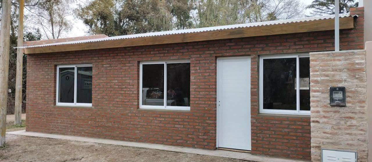 Edificio de ladrillo de una sola planta con tres ventanas, una puerta blanca y un pequeño letrero en el lado derecho.