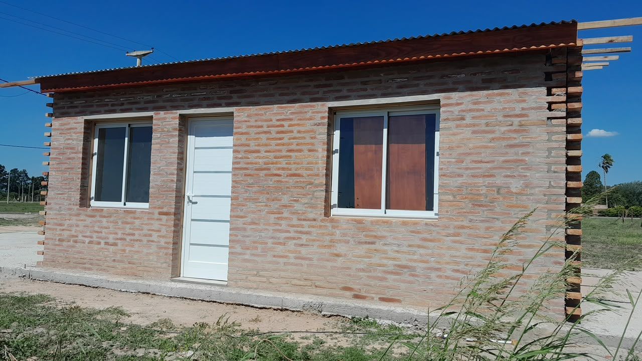 Pequeña casa de ladrillo con puerta blanca y dos ventanas bajo un tejado rojo en un entorno rural.
