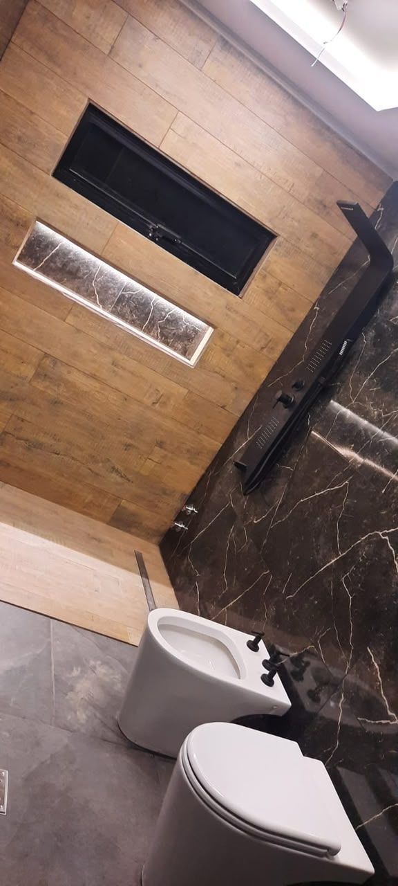 Baño pequeño con dos inodoros, suelo de baldosas y una pared de madera con una estrecha abertura para la ventana.