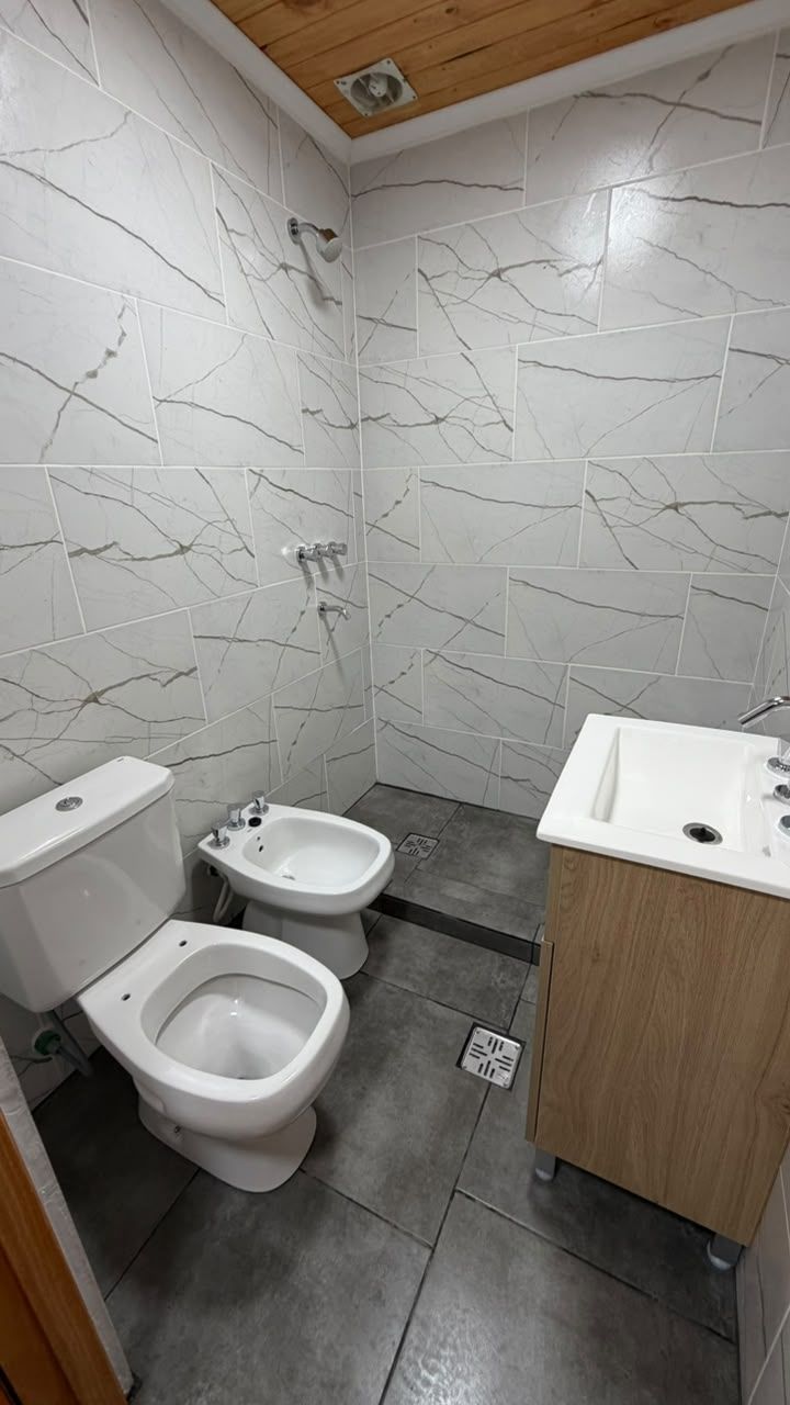 Baño pequeño y moderno con inodoro, bidé, mueble de lavabo, suelo de baldosas grises y paredes blancas texturizadas.