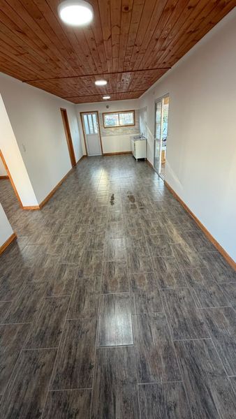 Pasillo estrecho y vacío con techo de madera, suelo gris, paredes blancas y ventanas al final.