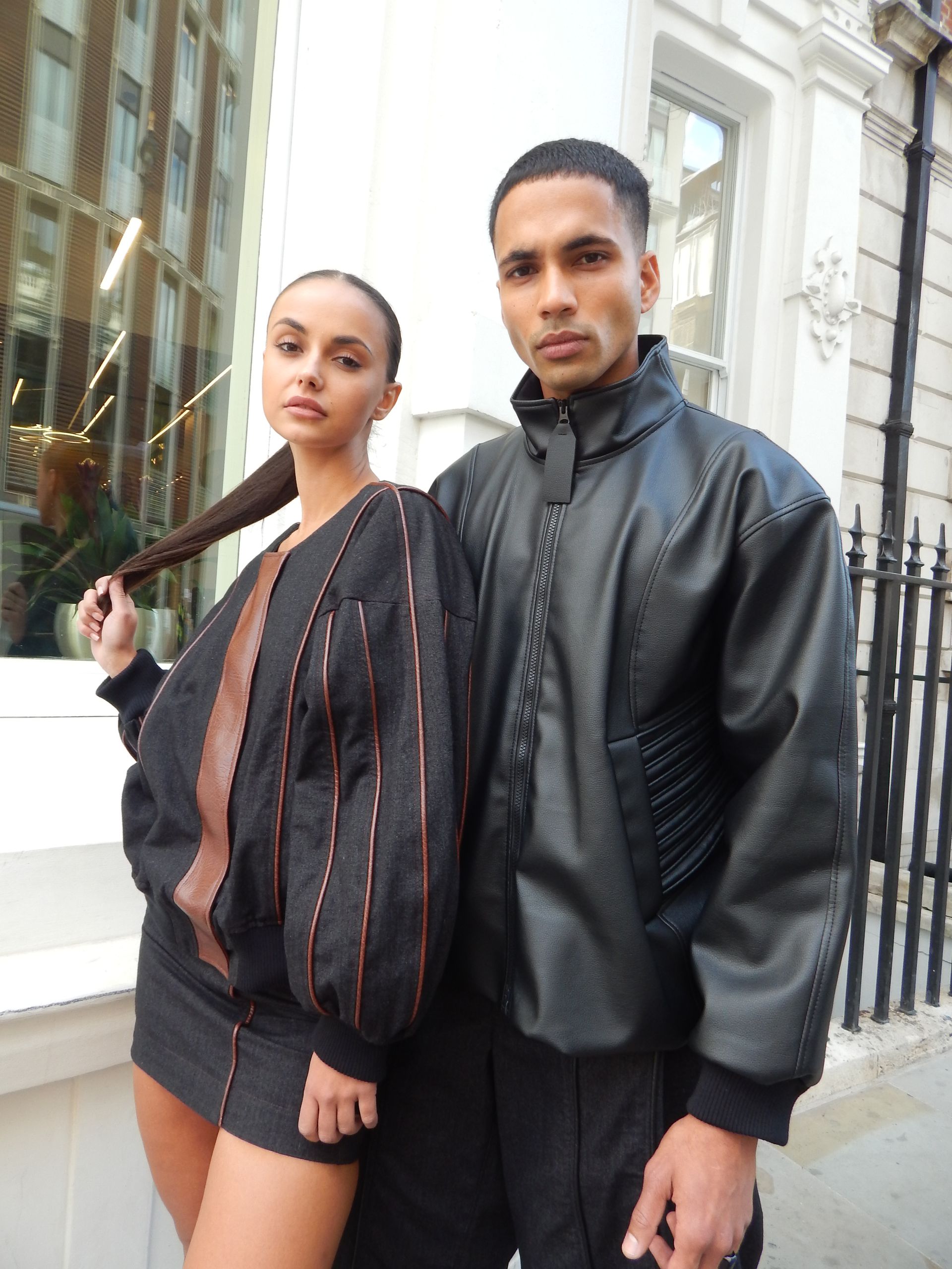 Couple in black outfits posing outside a building. The woman holds her braid, the man looks forward.