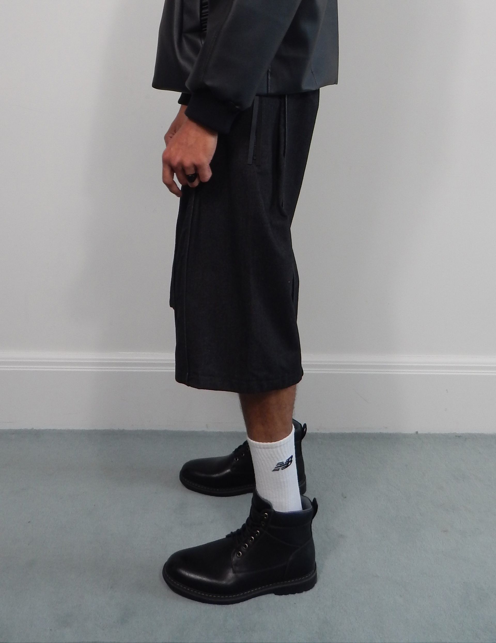 Person wearing black shorts, jacket, white socks, and black shoes, standing against a white wall.