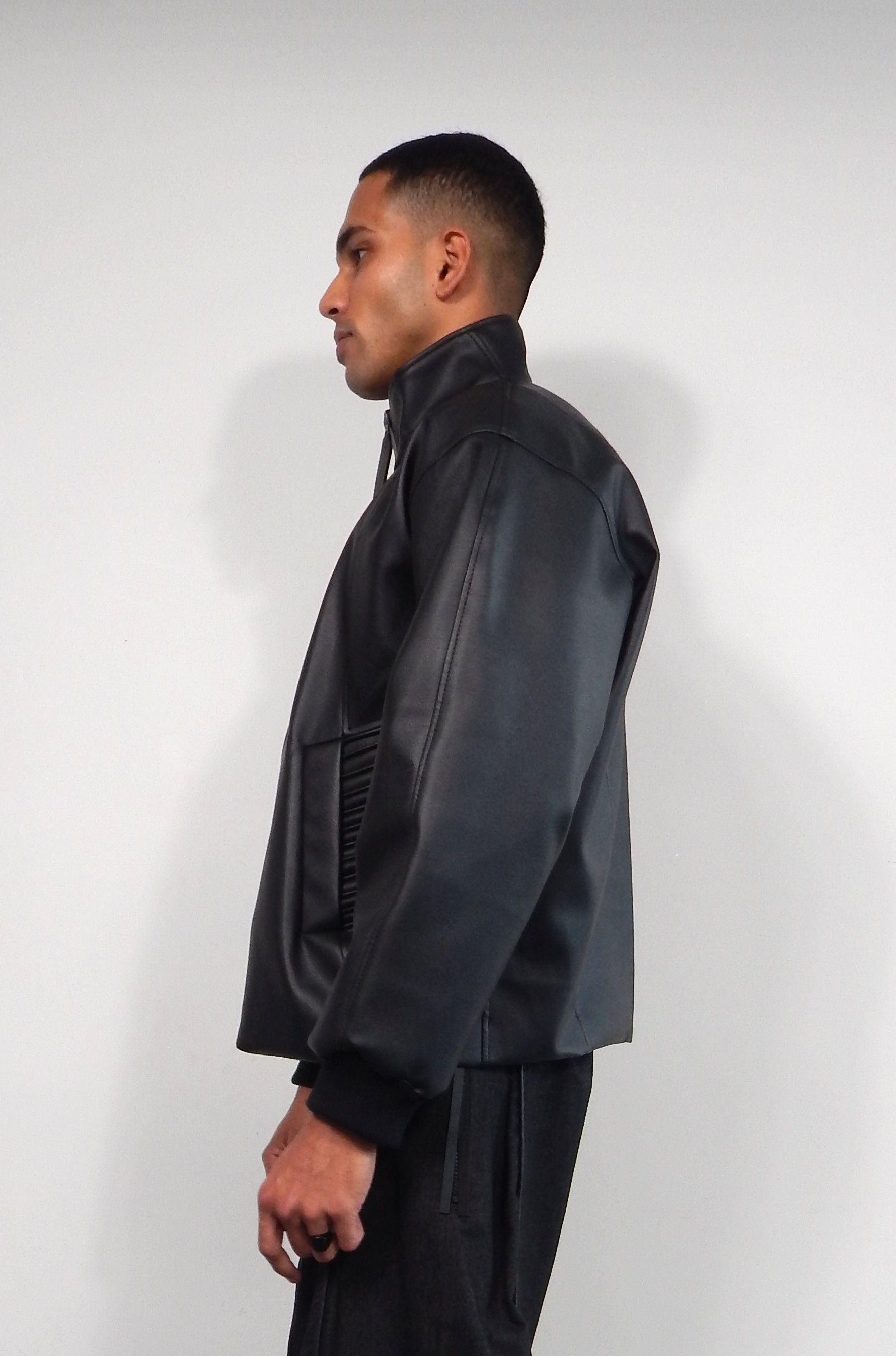 Man wearing a black jacket and black pants, standing in front of a white wall.