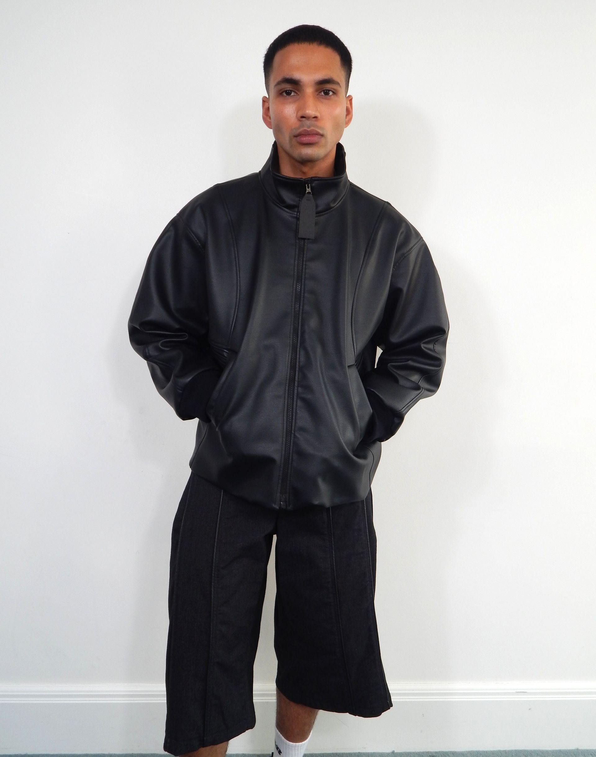 Person in black jacket and shorts stands against a white wall.