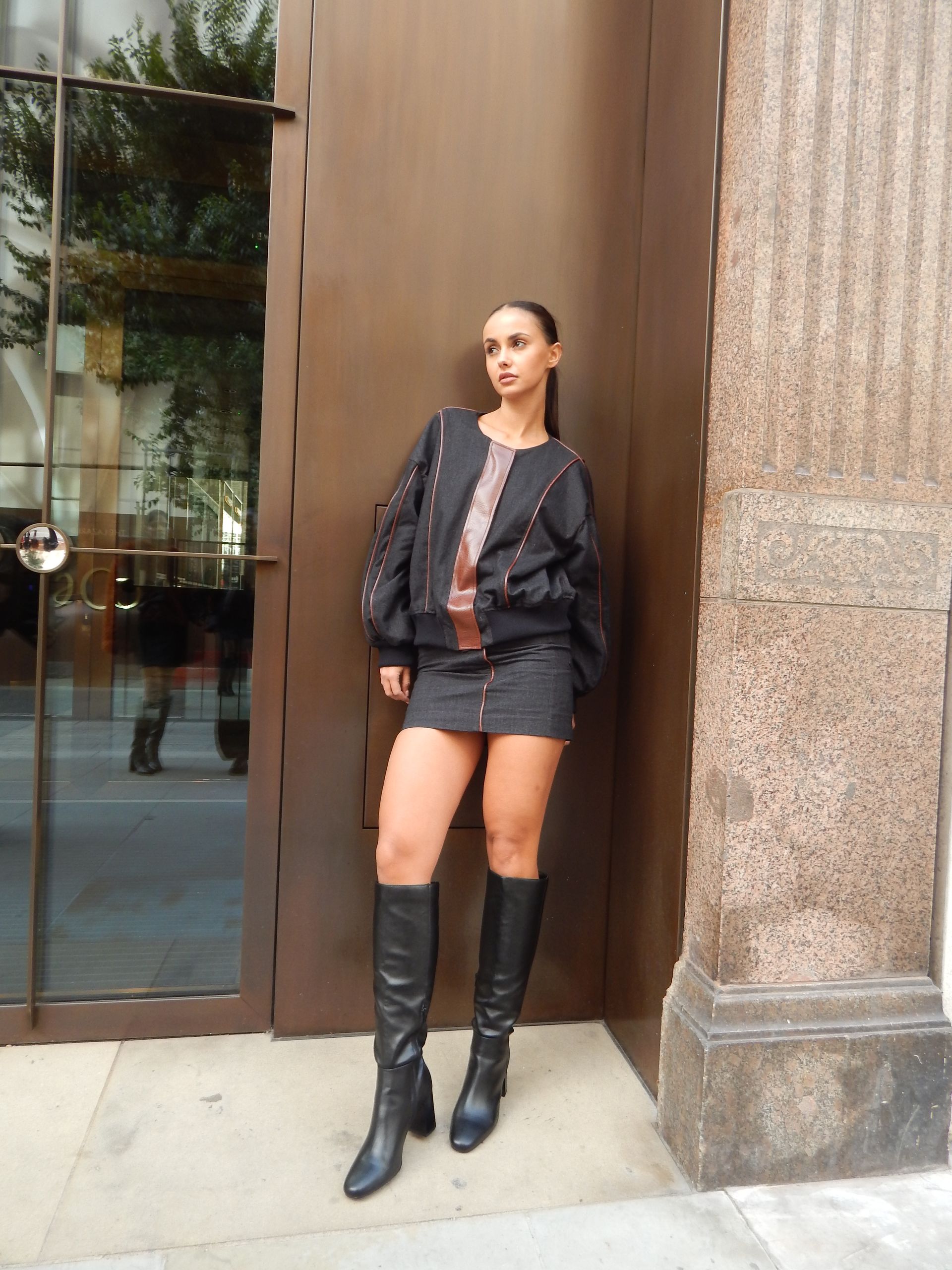 Woman in black bomber jacket, skirt, and knee-high boots, standing by a brown building.