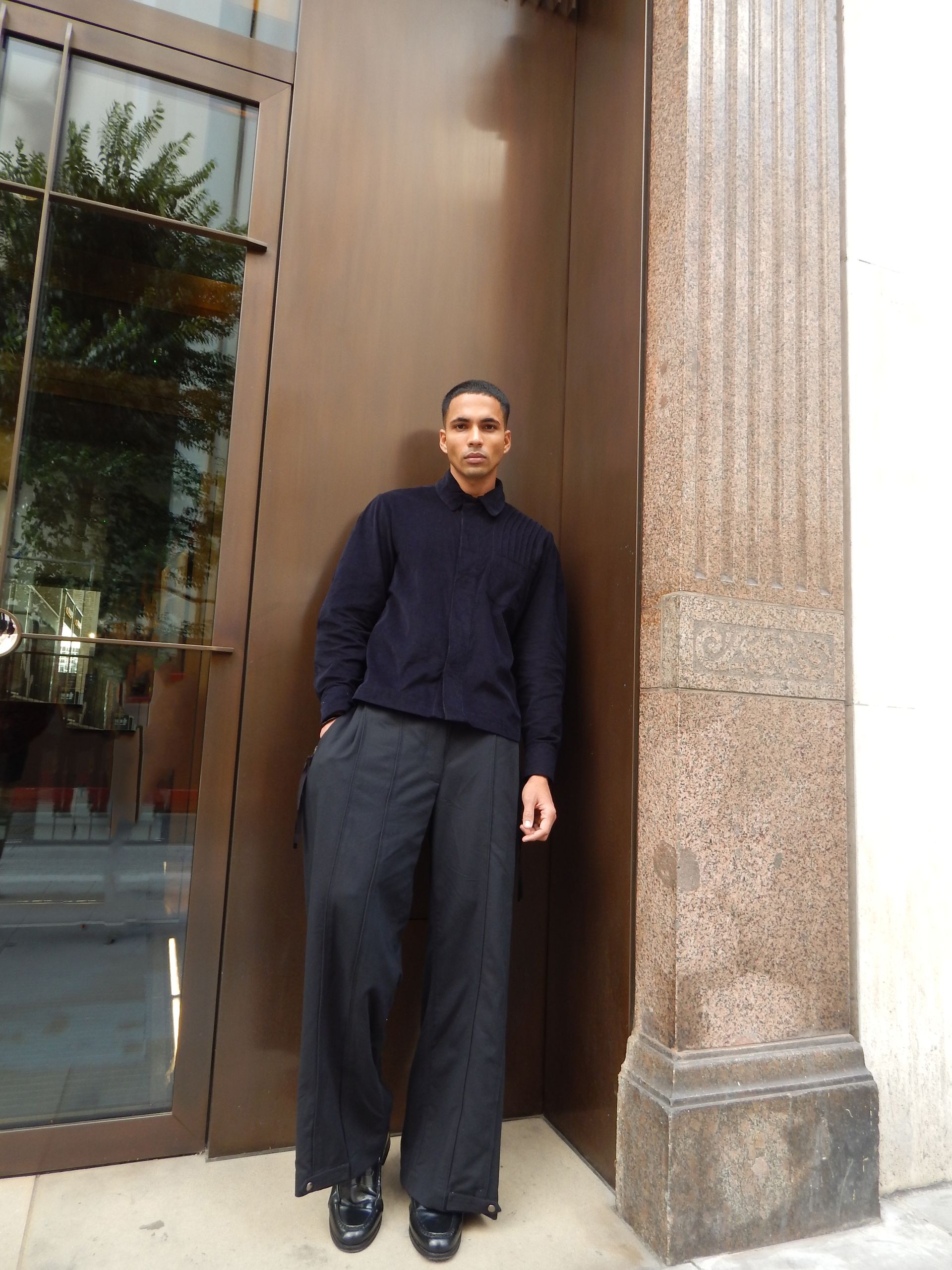 Man wearing dark sweater and wide-leg pants stands next to brown door and column.