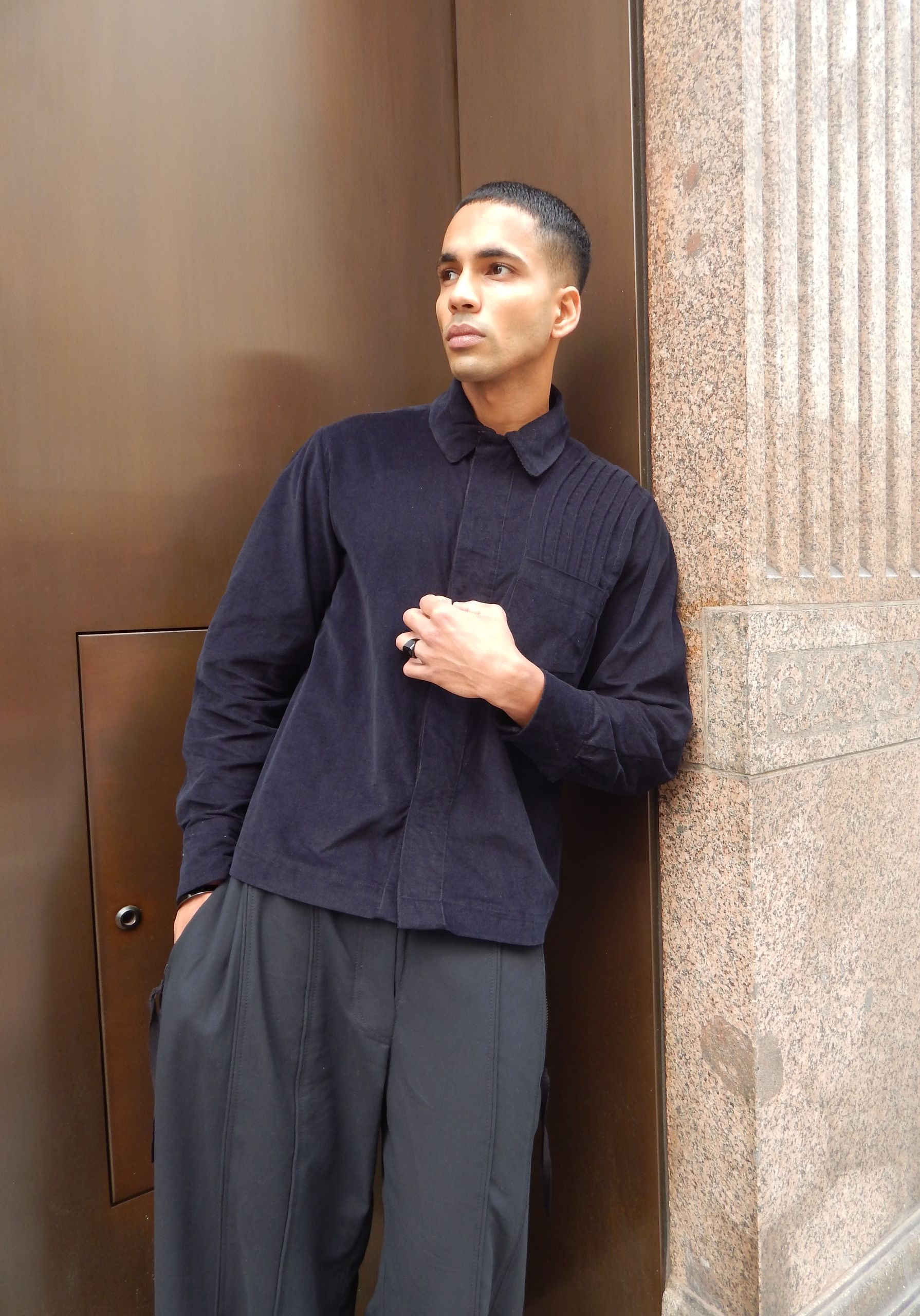 Man in dark shirt and pants leaning against a brown door and textured column, looking to the side.