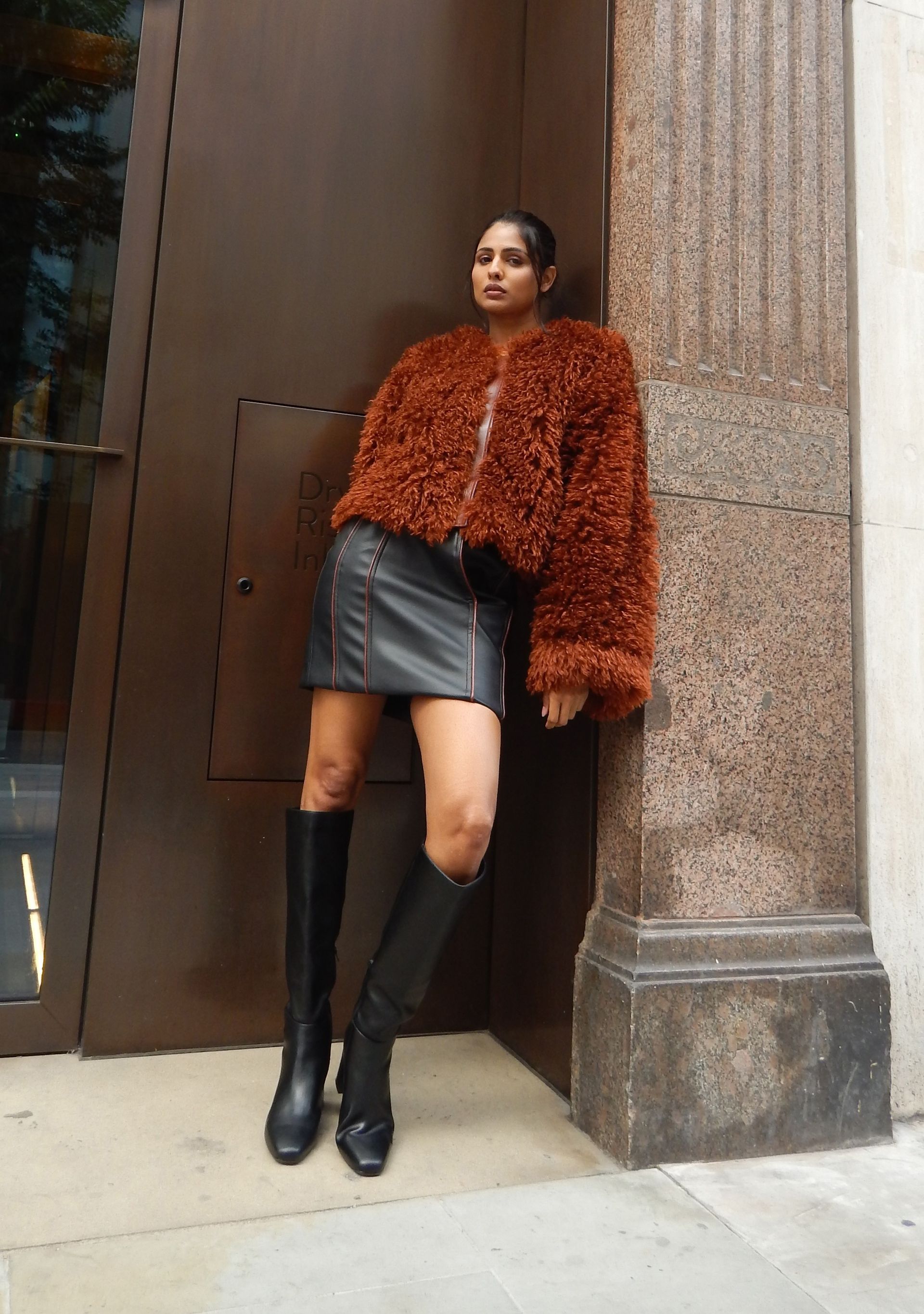 Woman in brown jacket, black skirt, and boots leans against a building.