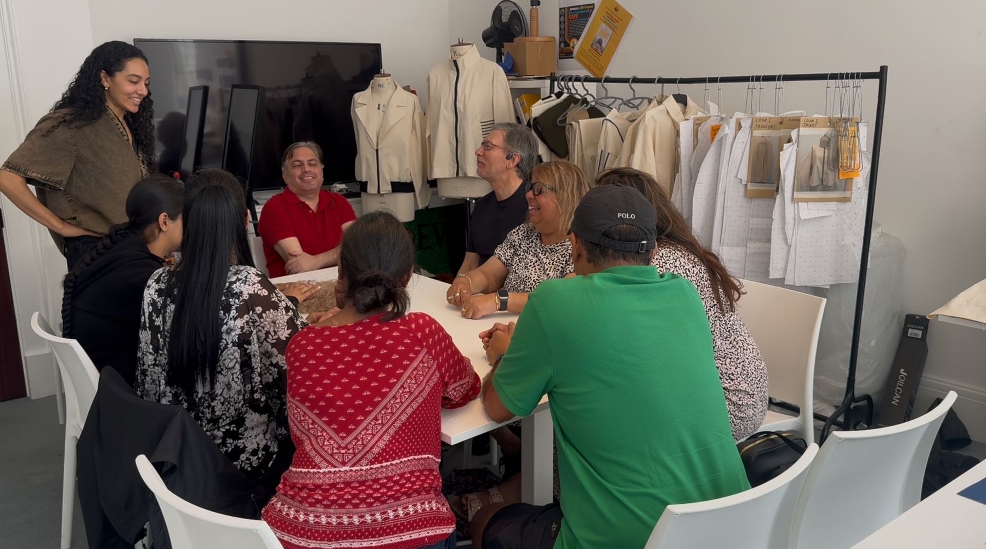 Group of people seated around a table, discussing. Some clothing hangs on a rack in the background.