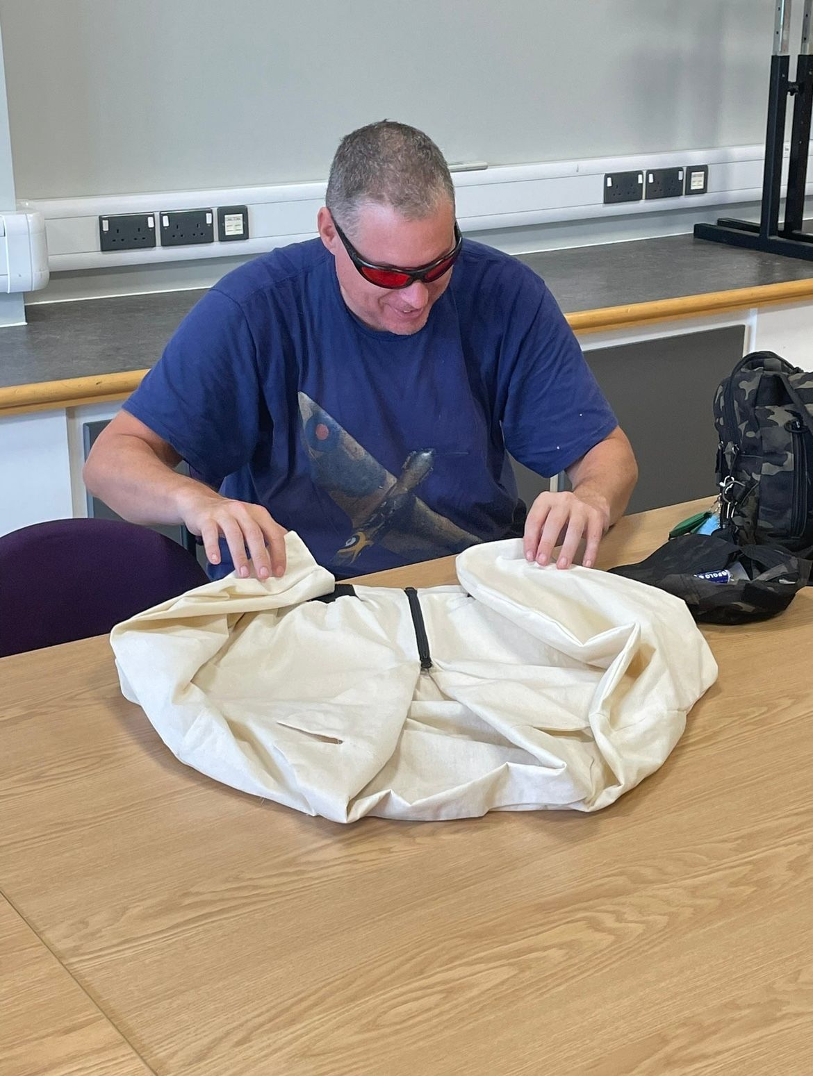 Man wearing sunglasses folding a light-colored jacket on a wooden table. Indoors with other items nearby.