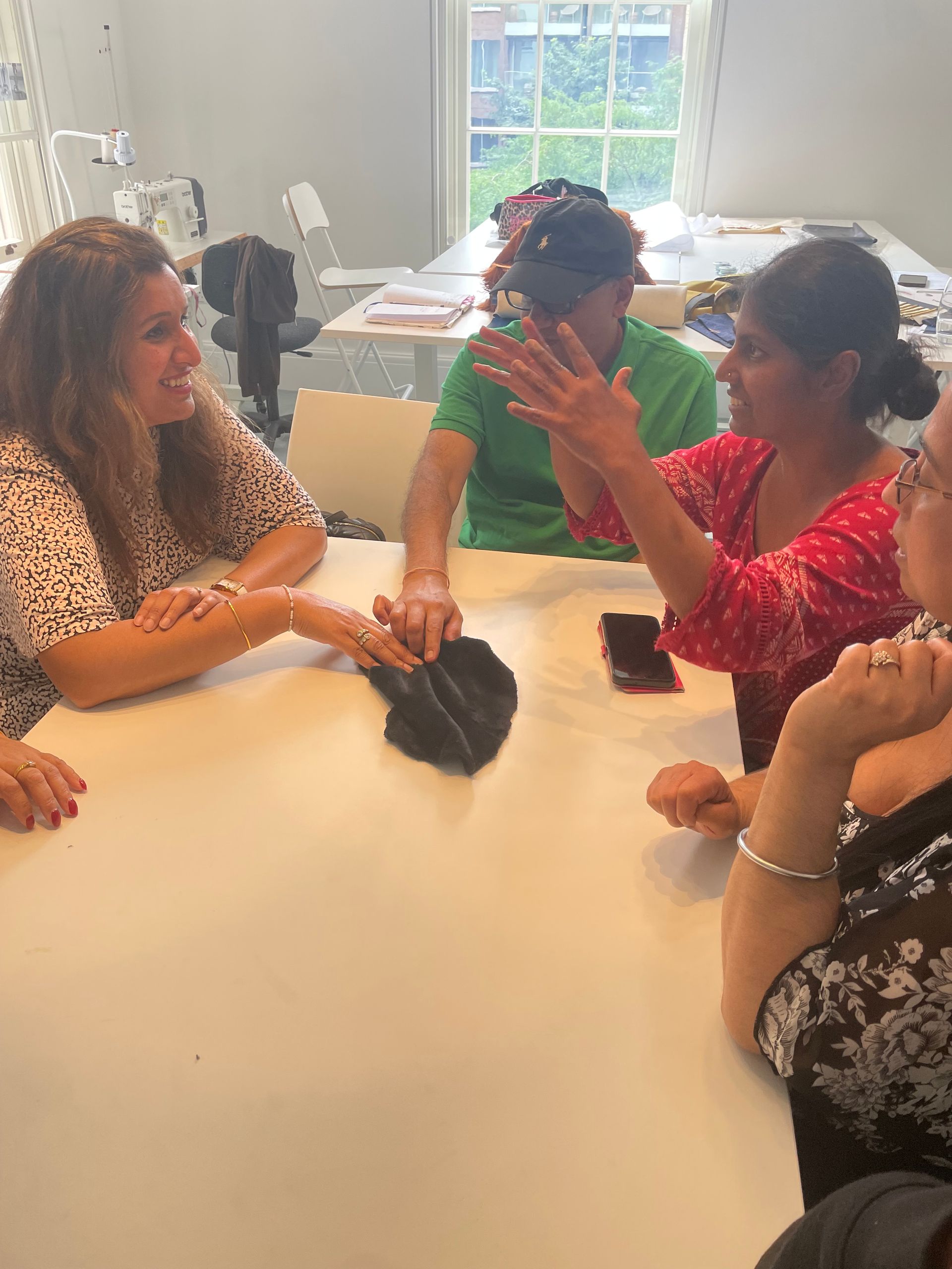 People seated around a table, discussing. Woman in red gestures. Black object in center. Bright room, natural light.