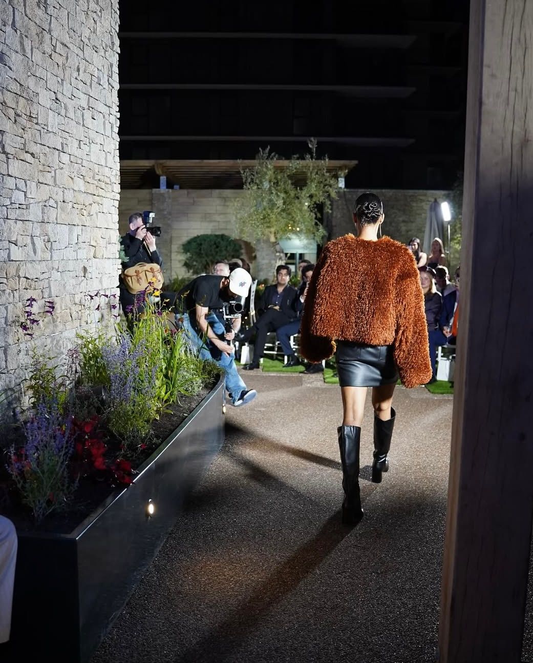 Fashion model in brown coat and black skirt walking a runway outdoors at night.