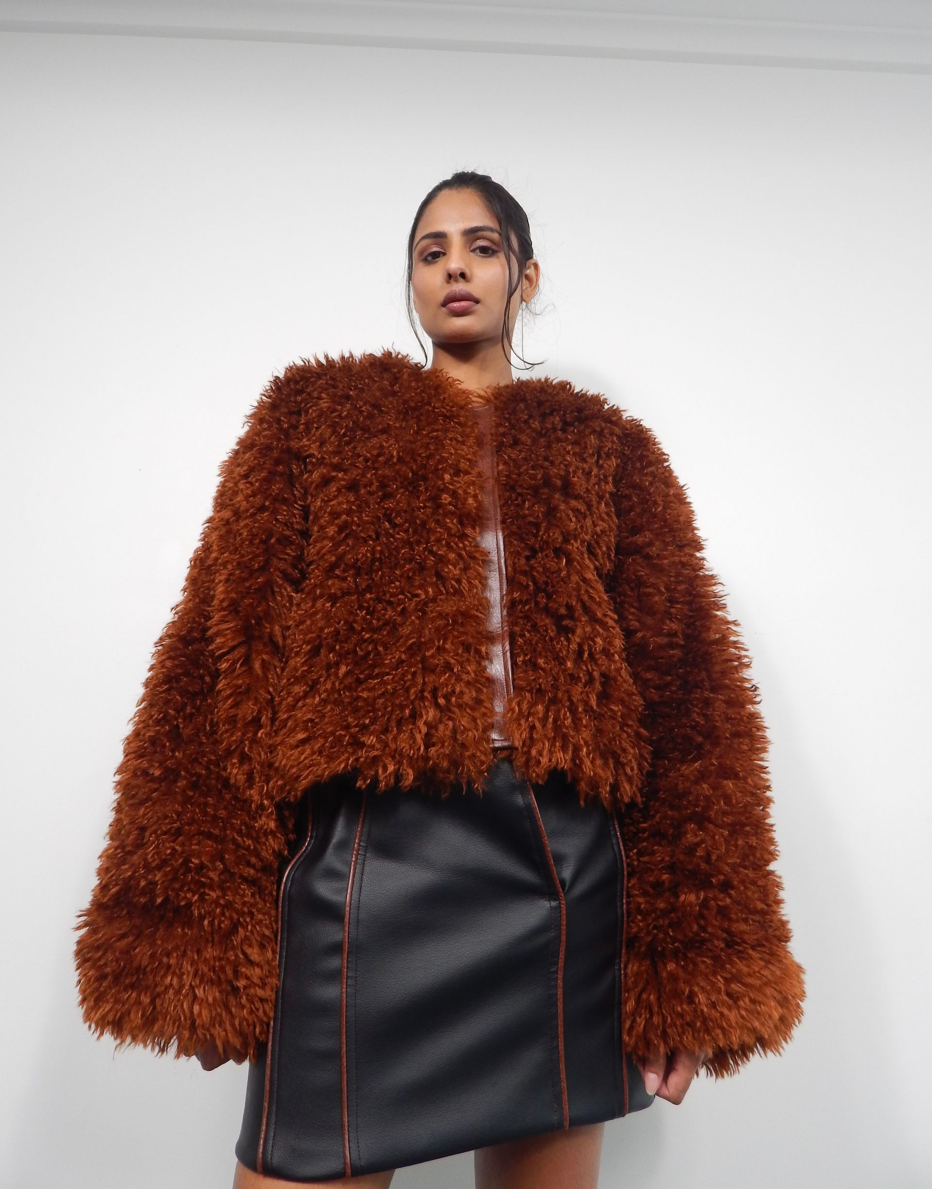 Woman wearing a brown furry jacket and black skirt. Facing forward, posing in a white setting.