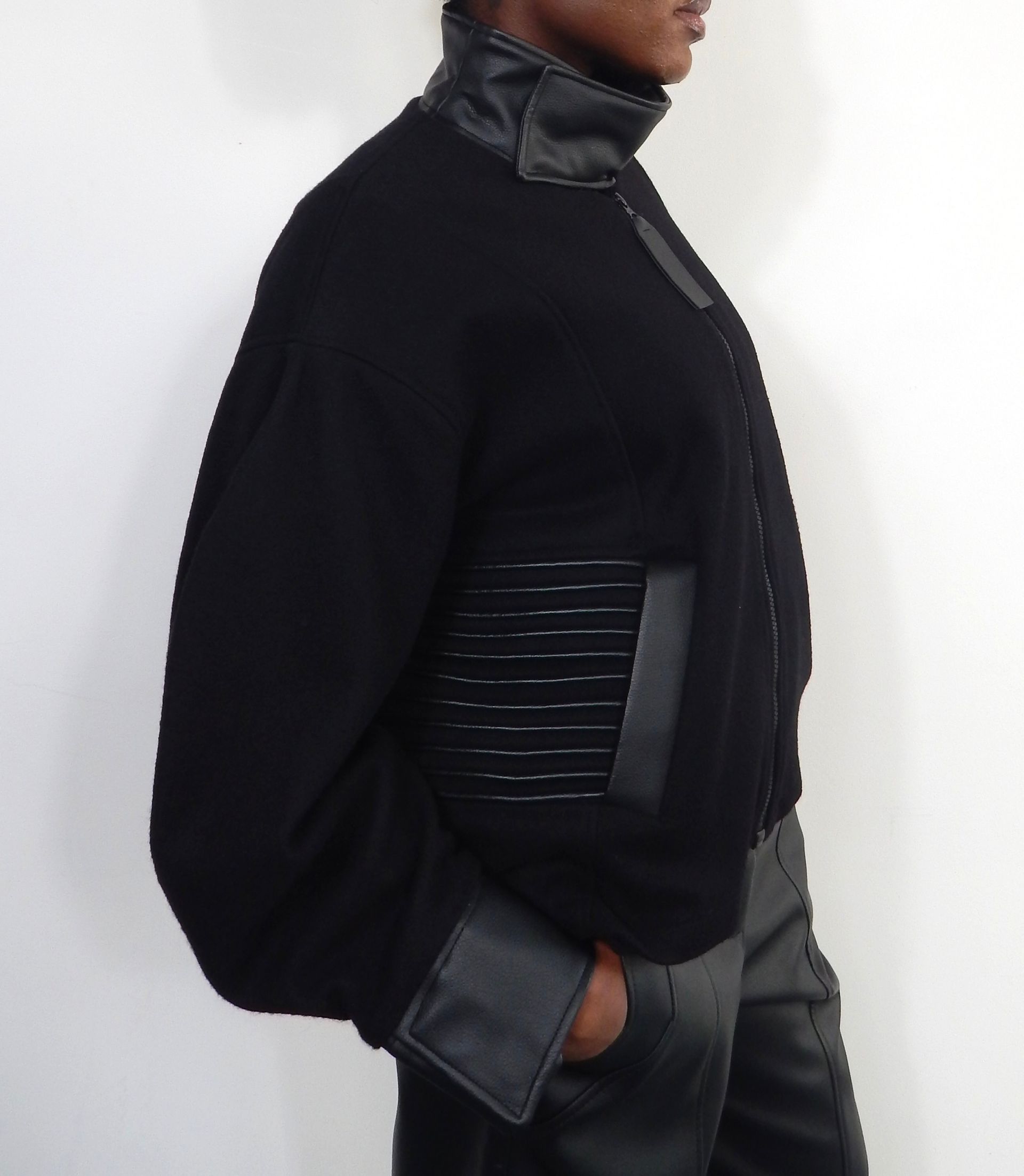 Person in black sweater with leather accents, standing. One hand in pocket.