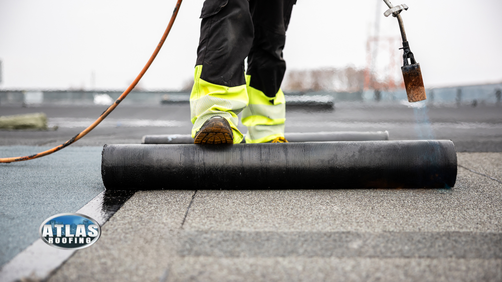 Worker spreading black roofing material on a rooftop with a torch and orange hose