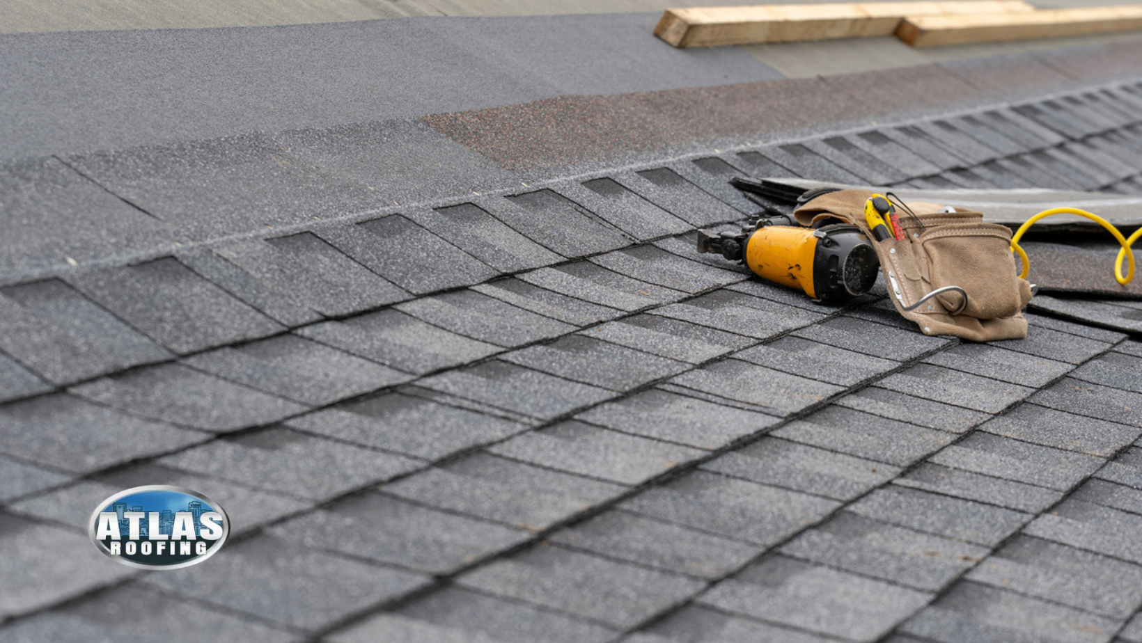 A close-up view of a dark gray shingle roof with a nail gun and tool belt resting on it.