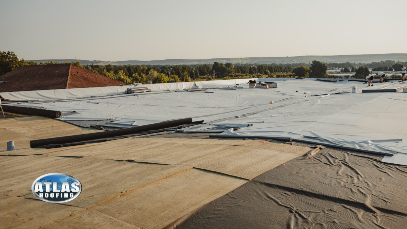 Rooftop with white membrane being installed, trees and buildings in background.