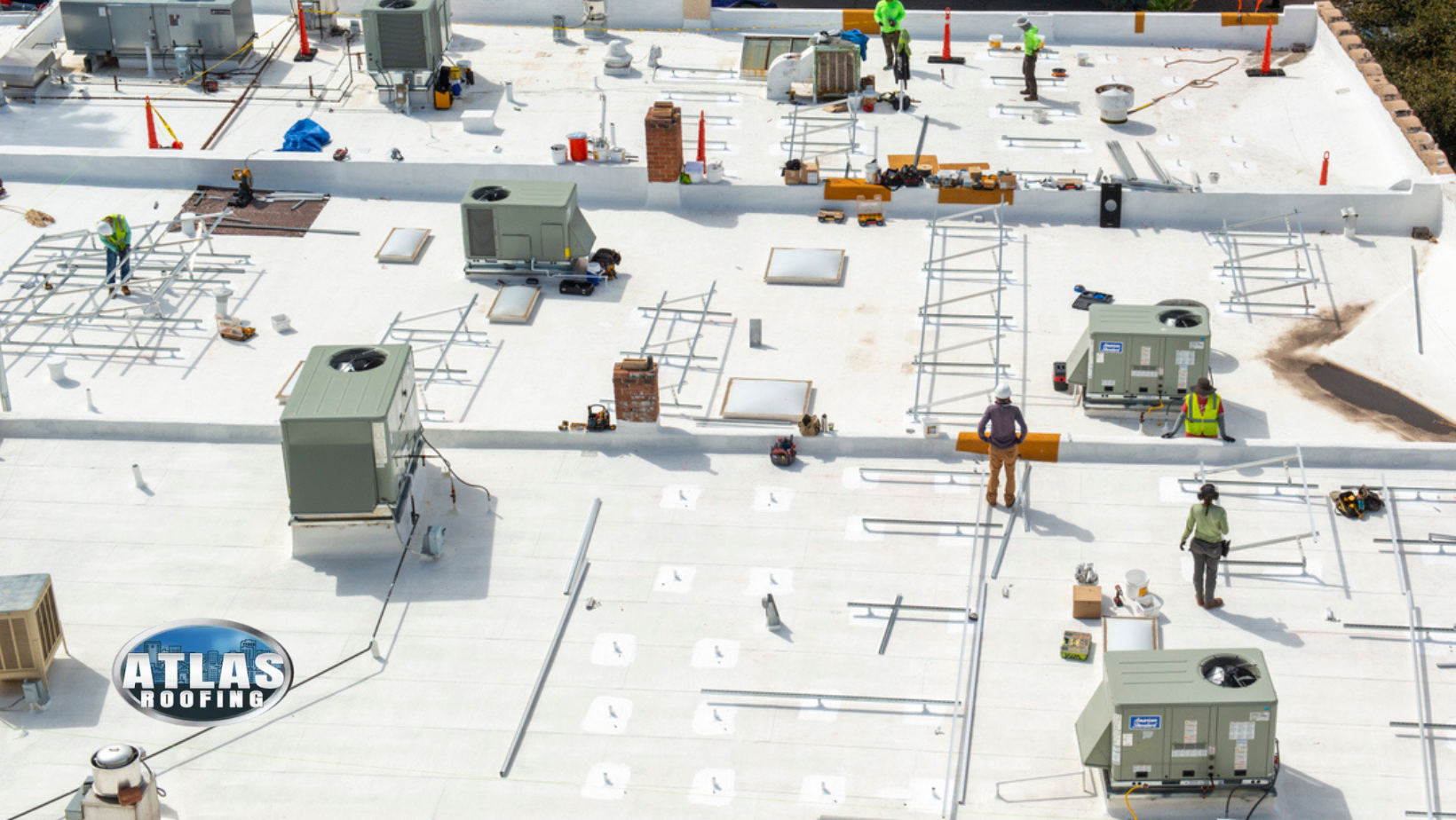 Workers on a rooftop install equipment. Gray HVAC units, white roof, safety cones.