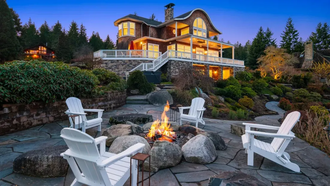 Cozy lodge at dusk with lit fire pit and white Adirondack chairs on a stone patio