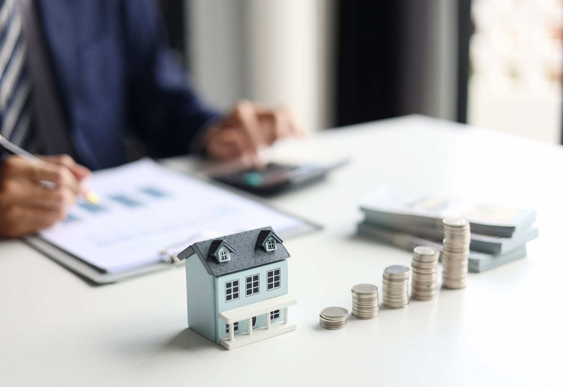 Miniature house with stacked coins on a desk, with a person reviewing paperwork in the background