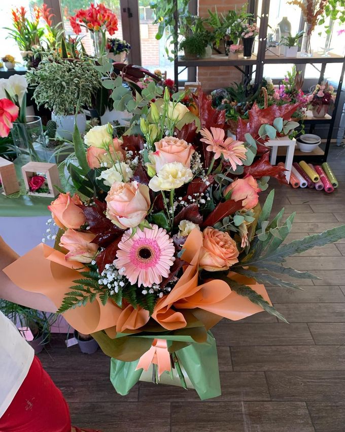 Bouquet di fiori misti color pesca e rosa con foglie verdi e confezione arancione, in un negozio di fiori.