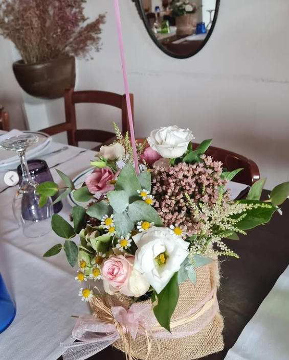 Centrotavola floreale con fiori rosa, bianchi e verdi in un vaso intrecciato su un tavolo da pranzo.